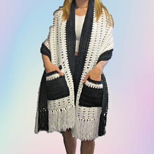 May include: A handmade crochet shawl in black and white. The shawl features a fringed hem and two front pockets. The black trim and pockets contrast with the white body of the shawl. The shawl is designed to be worn as a wrap.