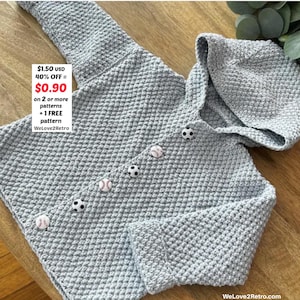 Puede incluir: Una chaqueta de bebé de crochet gris claro y texturizado con capucha y botones de béisbol y fútbol. La chaqueta tiene mangas largas y se muestra sobre una superficie de madera. El texto "WeLove2Retro" es visible.