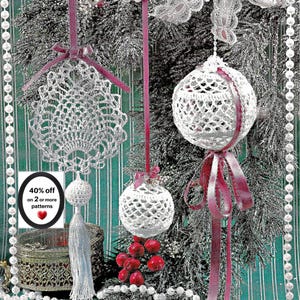 May include: Three white crocheted ornaments hanging from dark pink ribbons. One is a flat, intricate lace shape with a tassel, and two are spherical, open-work designs. A small sign reads 40% off on 2 or more patterns with a red heart icon.