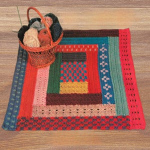 Può includere: Una colorata coperta all'uncinetto fatta a mano con un design geometrico. La coperta presenta una varietà di colori, tra cui rosso, blu, verde e giallo. Un piccolo cestino di vimini pieno di gomitoli di lana è posto sulla coperta. La coperta è su una superficie di legno.
