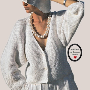 May include: A white knit cardigan with long sleeves and a V-neckline, featuring two visible buttons down the front. A large pearl-style beaded necklace is worn around the neck. A black oval graphic contains the text 40% off on 2 or more patterns.