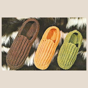 May include: Three slip-on slippers featuring a thick, ribbed crochet texture. The slippers are arranged in a row, displaying three distinct colors: a dark brown, a warm golden yellow, and a muted olive green, each with a rounded toe design.