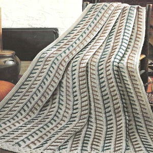 May include: A cream-colored crocheted blanket with a repeating pattern of vertical stripes. Each stripe features a series of alternating brown and teal-colored stitches. The blanket is draped over a chair, showcasing its texture and design.