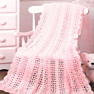 May include: A light pink, crocheted baby blanket draped over a white rocking chair. The blanket has a delicate, openwork design with a scalloped edge. A matching pink teddy bear sits nearby, suggesting a nursery setting.