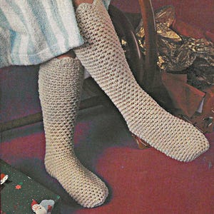 Könnte beinhalten: Ein Paar beige, kniehohe Häkelsocken. Die Socken sind mit einem strukturierten Stich gefertigt und scheinen handgefertigt zu sein. Der Hintergrund umfasst eine rote Oberfläche und ein gestreiftes Handtuch.