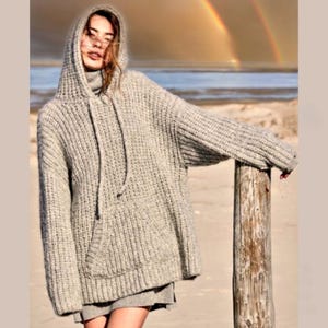 Pode incluir: Um moletom cinza de malha oversized com um bolso canguru e cordão. O suéter de malha grossa tem capuz e é usado com uma gola alta e saia cinza. O fundo apresenta uma cena de praia com um arco-íris.