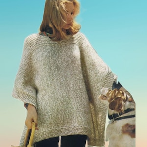 May include: A light beige knitted poncho with a wide neckline and short sleeves. The poncho has a textured appearance and a relaxed fit. The person is holding a basket. A dog is next to the person.