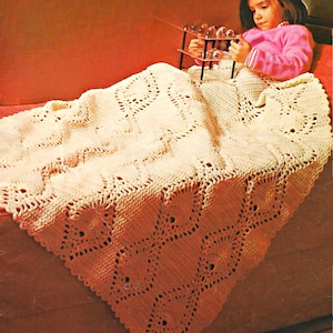 May include: A cream-colored crochet blanket featuring an intricate, repeating diamond-shaped pattern with openwork detailing. The textile has a textured, knotted surface and scalloped edges, draped over a surface with a soft, woven appearance.