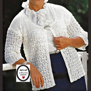 May include: A white crochet cardigan featuring a textured openwork pattern, three-quarter length sleeves, and a single decorative silver-toned button closure at the neckline. The garment is worn over a white ruffled blouse and dark blue trousers.