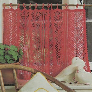 May include: A red, open-weave curtain hangs in front of a window. The curtain has a decorative, geometric pattern. A potted plant and two white ceramic doves are visible in front of the curtain.