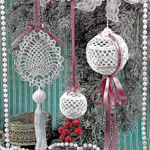 Puede incluir: Adornos navideños blancos de ganchillo con cintas rosas colgados de un árbol decorado. Los adornos incluyen una bola redonda, un diseño en forma de abanico y una bola más pequeña. También se ven bayas rojas y una caja decorativa.
