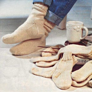 May include: Cream-colored knitted socks, one pair worn and another pair nearby. The socks are ribbed and appear handmade. A white mug and knitting supplies are also visible, suggesting a cozy, craft-focused theme.