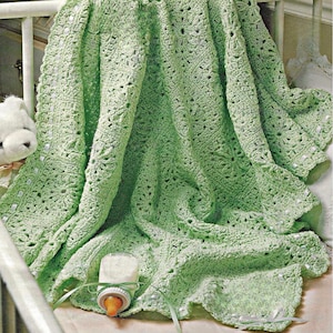 May include: A light green crocheted baby blanket with a floral pattern is draped over a crib. A baby bottle with a white bottle and orange nipple is in the foreground. A white teddy bear is visible on the left side of the image.