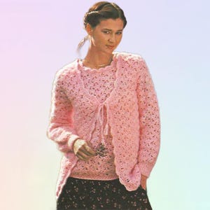 May include: A light pink crochet cardigan and matching top. The open-front cardigan has long sleeves and a tie closure. The top has a round neckline. The crochet pattern is open and airy. The outfit is worn over a black floral skirt.