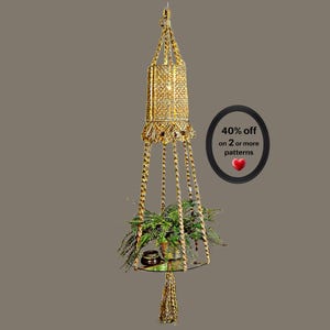 May include: A gold-colored macrame plant hanger featuring a cylindrical top section, braided support cords, and a circular glass shelf holding a green fern. The text 40% off on 2 or more patterns with a red heart icon is displayed to the right.