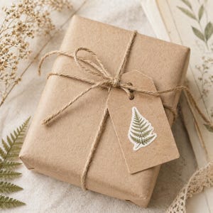 May include: A wrapped gift in brown paper, tied with twine and a bow. A small, rectangular tag with a fern illustration is attached. The gift is on a neutral-colored surface, with dried plants and lace trim in the background.