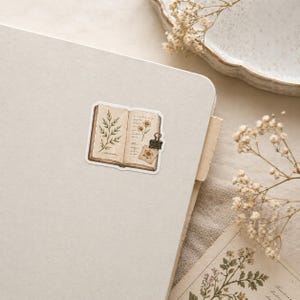 May include: A sticker featuring an open book illustration with botanical drawings and a small paper clipped to it. The sticker has a vintage aesthetic with sepia tones and is placed on a light-colored surface.