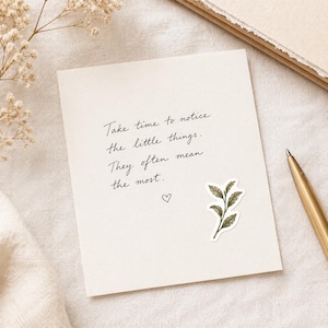 May include: A cream-colored card with handwritten text: "Take time to notice the little things. They often mean the most." A small green and brown leaf sticker and a heart are also on the card. A gold pen rests on the right.