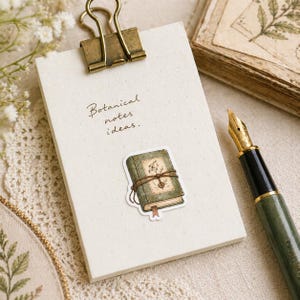 May include: A cream-colored clipboard with a brass clip holds a notepad with the text "Botanical notes ideas." A sticker of a vintage book is attached. A green and gold fountain pen rests nearby, suggesting a stationery theme.