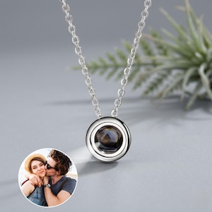 Op de afbeelding: Een zilverkleurige hangerketting met een ronde, heldere kern. De ketting is ook zilver. Een kleine, cirkelvormige afbeelding van een stel staat in de linkerbenedenhoek. De achtergrond is lichtgrijs.