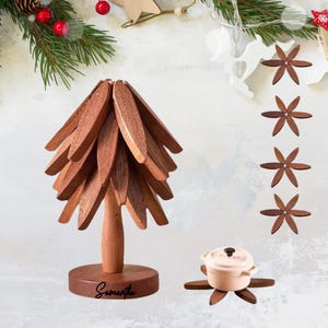 May include: A wooden Christmas tree-shaped decoration with a round base, and the name "Samantha" engraved on the base. A small, light pink pot sits on a wooden trivet. Additional wooden star-shaped ornaments and festive greenery complete the holiday theme.