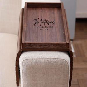 May include: A dark brown wooden sofa arm tray with a rectangular surface and raised edges. The tray is personalized with the text "The Petersons", "OLIVIA & SPENCER", and "EST. 2026". The tray is placed on a beige upholstered sofa arm.