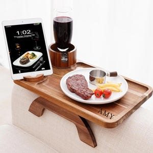 May include: A wooden sofa arm tray with a built-in drink holder and tablet stand. A plate with steak, fries, and tomatoes sits on the tray. A glass of red wine and a tablet displaying a meal are also present. The tray is engraved with the name "AMY".