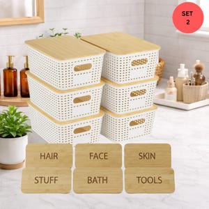 May include: A set of six white plastic storage bins with bamboo lids. Each bin has a rectangular shape with a perforated design. Six bamboo labels are included, with the words "HAIR", "FACE", "SKIN", "STUFF", "BATH", and "TOOLS" engraved on them.
