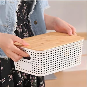 May include: A white rectangular storage basket with a bamboo lid. The basket has a perforated design and integrated handles. The lid is a light brown color. The basket is being held by a person.