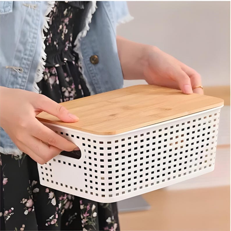 May include: A white rectangular storage basket with a bamboo lid. The basket has a perforated design and integrated handles. The lid is a light brown color. The basket is being held by a person.