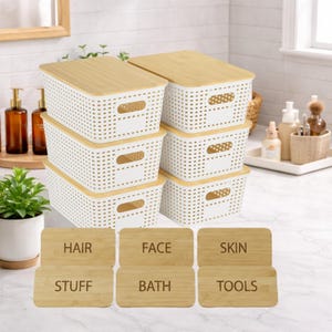 May include: A set of six white plastic storage bins with bamboo lids. Each bin has a rectangular shape with a perforated design. The labels read "HAIR", "FACE", "SKIN", "STUFF", "BATH", and "TOOLS".