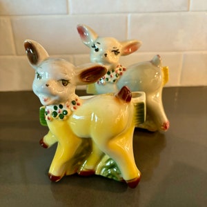 May include: Two ceramic deer planter figurines. The deer on the left is yellow with green and red accents. The deer on the right is blue with green and red accents. Both deer have painted eyes and a floral collar.