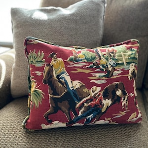May include: A rectangular throw pillow featuring a red fabric cover with a printed pattern of cowboys riding horses and bucking bulls. The design includes figures in yellow shirts and blue jeans, with green foliage accents and a piped edge finish.