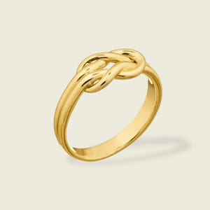 May include: A gold ring with a knot design. The ring has a polished finish and a classic, elegant style. The knot is the focal point, with the band curving around to form the ring's structure. A timeless piece of jewellery.