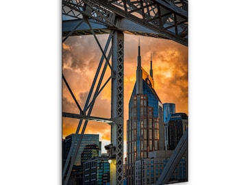 Puede incluir: Una impresión de la ciudad de Nashville, Tennessee, enmarcada por una estructura de puente. La imagen captura el horizonte de la ciudad contra una vibrante puesta de sol, con el icónico edificio AT&T en primer plano.