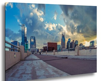 Puede incluir: Una impresión de paisaje urbano con un horizonte moderno bajo un cielo dramático. La imagen muestra un puente que conduce a la ciudad, con edificios altos y rascacielos. El cielo está lleno de nubes, creando una escena dinámica y colorida.