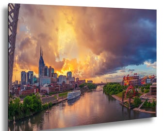 Puede incluir: Impresión en lienzo de un paisaje urbano al atardecer. La imagen presenta un río, edificios y un cielo dramático con tonos naranja, amarillo y púrpura. También se ven un puente y un barco.