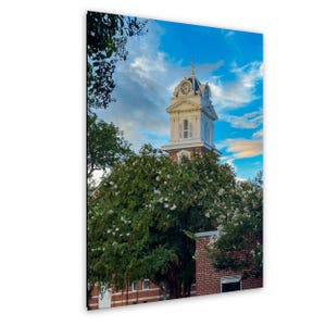 May include: A vertical print of a historic building with a clock tower. The tower is white with a clock face and a cross on top. The building has a red brick base and is surrounded by green trees and a blue sky with clouds.