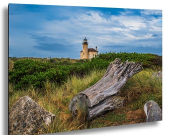 Pode incluir: Uma fotografia de paisagem com um tronco de madeira flutuante cinzento e desgastado em ervas costeiras verde-douradas. Ao fundo, um farol de cor clara ergue-se contra um céu azul nublado sobre um denso arbusto verde.