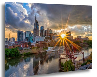 Puede incluir: Una impresión en lienzo del horizonte de Nashville al atardecer. La imagen captura los edificios de la ciudad, un río y un cielo dramático con rayos de sol. Un barco está atracado en el río. La paleta de colores incluye tonos de naranja, azul y gris.