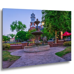 May include: A stone tiered fountain sits in a park plaza with a paved walkway in the foreground. In the background, a multi-story red brick building with a white clock tower stands among green trees under a clear twilight sky with street lamps.