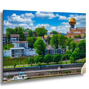 Puede incluir: Una vista panorámica de una ciudad con una estructura dorada en forma de esfera sobre un edificio. Frondosos árboles verdes y edificios de diversos estilos arquitectónicos se alzan bajo un cielo azul nublado. Un tren recorre la orilla del agua.