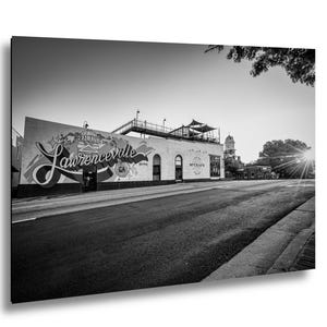 May include: A black and white photograph of a street scene in Lawrenceville, Georgia. A large building features a mural with the text 'It's a beautiful day in Lawrenceville GA' and 'McCray's Tavern'. A rooftop deck is visible above the building.
