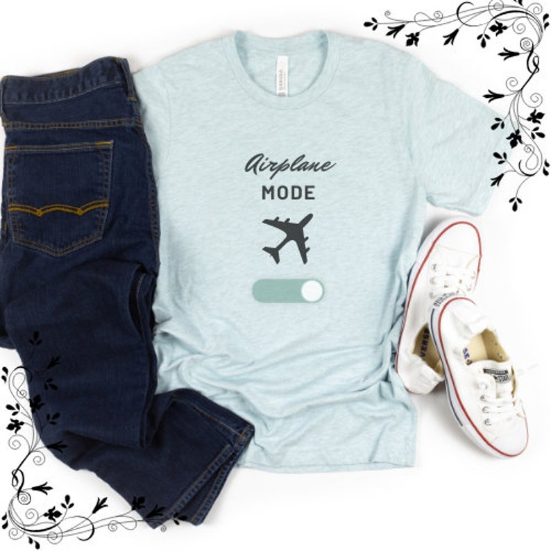 May include: A pale blue t-shirt featuring the text "Airplane Mode" and an airplane graphic with a toggle switch. The shirt is styled with dark blue jeans and white trainers.