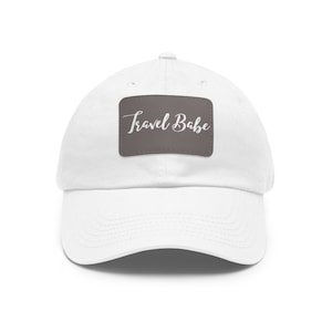 Op de afbeelding: Witte baseballpet met een bruin patch waarop in witte cursieve letters "Travel Babe" staat.
