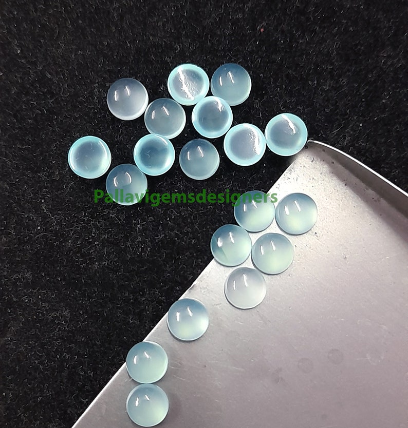 May include: A collection of pale blue cabochon gemstones, some on a silver tray and some scattered on a black surface.