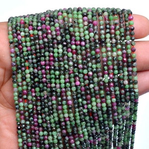 Pode incluir: Um fio de contas de pedras preciosas facetadas verdes, rosa e pretas. As contas são pequenas e redondas, e são enfiadas em um fio fino.