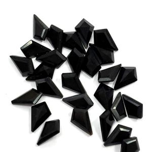 May include: A scattering of black gemstone beads, each with a geometric design. The beads are faceted, catching the light, and are arranged on a white surface. The shapes include diamond and trapezoid forms.