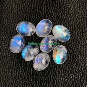 Puede incluir: Colección de piedras preciosas de forma ovalada, talladas en facetas. Las piedras lunares muestran una base blanca lechosa con destellos iridiscentes azules y amarillos. Las piedras están dispuestas sobre una superficie oscura y texturizada.