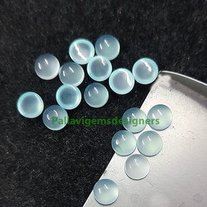 May include: A collection of 15 light blue round cabochon gemstones. The gemstones are smooth and polished, with a slightly translucent appearance. The gemstones are arranged in a loose pile on a black surface.
